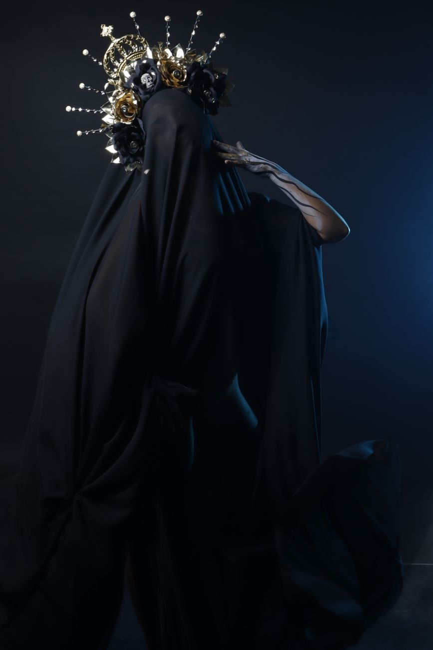 A mysterious figure draped in black with an ornate headdress in a dark, moody setting